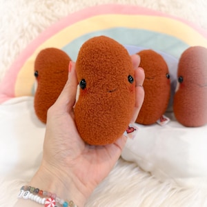May include: A hand holds a plush bean-shaped toy with a smiling face and black eyes. Several similar toys are in the background, along with a rainbow-shaped pillow. The toys are a reddish-brown color.