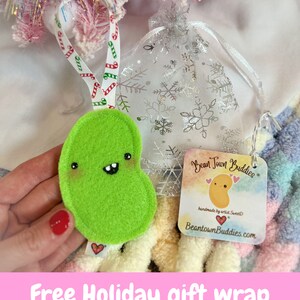 May include: A lime green felt bean-shaped ornament with a smiling face and a red heart tag. It hangs from a white ribbon with red and green candy cane designs. A silver mesh gift bag with snowflake patterns is also visible.