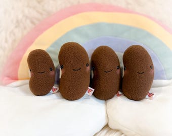 Puede incluir: Cuatro juguetes de peluche con forma de frijol marrón con caras sonrientes y pequeñas etiquetas de corazón, dispuestos sobre una almohada blanca con forma de nube. Un arcoíris pastel de peluche está en el fondo. Los juguetes tienen un aspecto suave y texturizado.