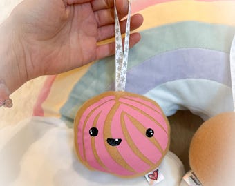 Può includere: Un ornamento di peluche rosa e marrone a forma di concha, con un volto sorridente e un nastro bianco con motivi a fiocchi di neve. L'ornamento è tenuto davanti a un cuscino arcobaleno.