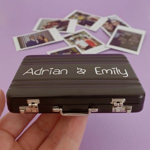 Op de afbeelding: Een zwarte metalen koffer met een zilveren handvat en sluitingen. De koffer is gegraveerd met de tekst "Adrian & Emily".