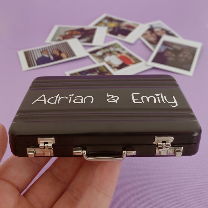 Op de afbeelding: Een zwarte metalen koffer met een zilveren handvat en sluitingen. De koffer is gegraveerd met de tekst "Adrian & Emily".