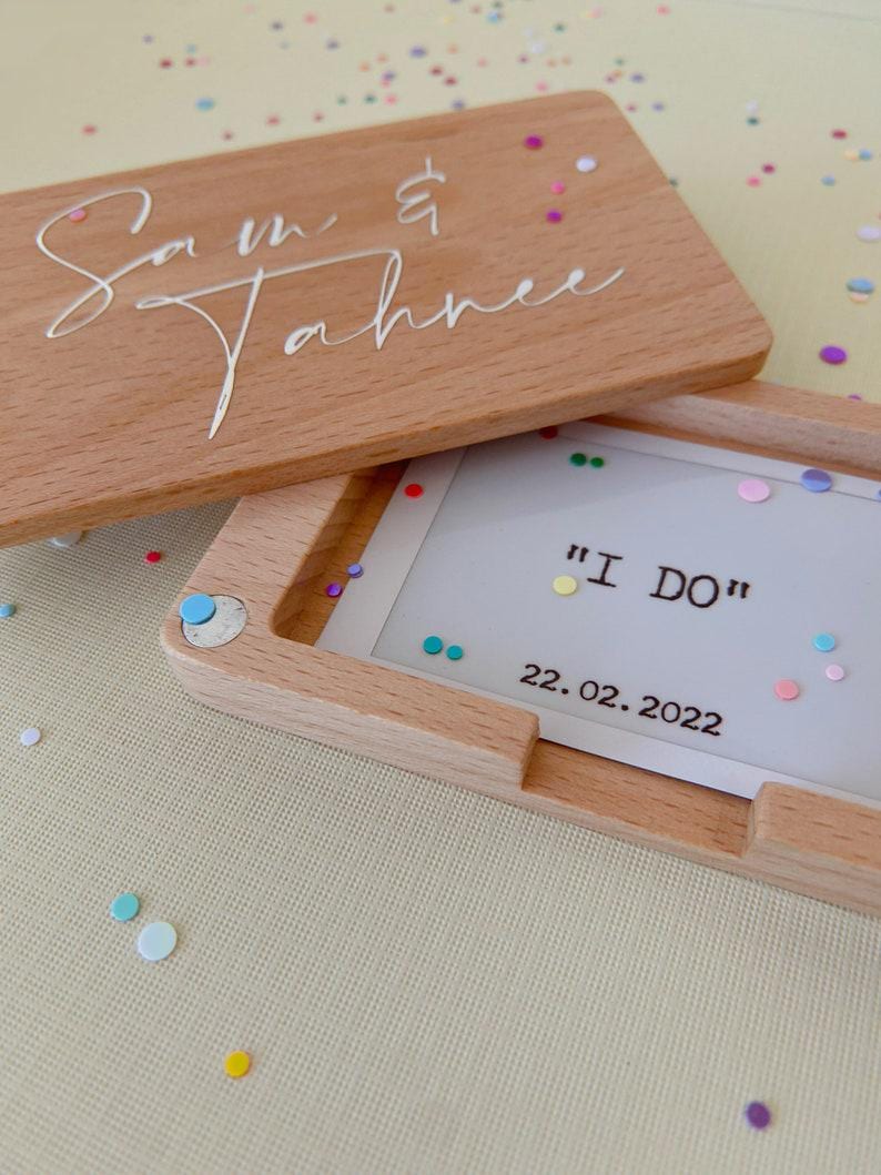 Op de afbeelding: Een houten doos met een deksel waarop "Sam & Tahnee" staat. De doos is open en er zit een witte kaart in met de woorden "I DO" en de datum "22.02.2022".