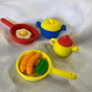 May include: Four colourful toy kitchen items: a red frying pan with a fried egg, a yellow pot with a blue lid, a yellow teapot with a red lid, and a yellow frying pan with sausages and green beans.