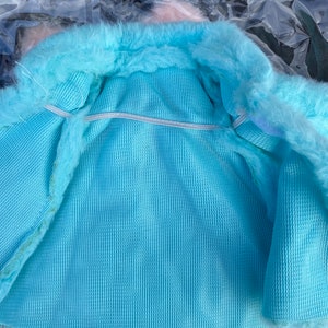 May include: A light blue faux fur jacket with a white elastic band around the waist. The jacket is lined with a light blue fabric.