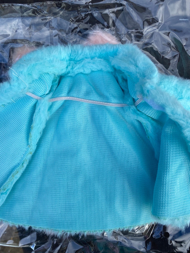 May include: A light blue faux fur jacket with a white elastic band around the waist. The jacket is lined with a light blue fabric.