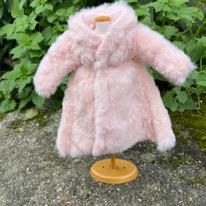 May include: A pink faux fur coat with a collar and long sleeves. The coat is displayed on a wooden mannequin.