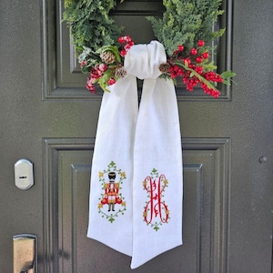 May include: A Christmas wreath with red berries, pinecones, and greenery hangs on a dark green door. Two white fabric panels are draped below the wreath, each embroidered with a festive design: a nutcracker and a monogram.