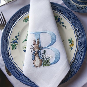May include: A white linen napkin with blue embroidery of a rabbit holding a blue letter B. The napkin is folded and placed on a blue and white patterned plate.