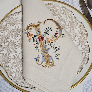 Könnte beinhalten: Eine gefaltete Leinen-Serviette mit einem gestickten Buchstaben "P" in Gold, Blau und floralen Akzenten. Die Serviette liegt auf einem goldgeränderten Teller mit floralem Muster, daneben ein Messer und ein Löffel. Thema: elegantes Essen.
