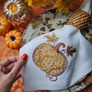 Puede incluir: Servilleta de lino blanco con un diseño de calabaza bordada en tonos dorados y marrones. La servilleta forma parte de una mesa temática otoñal, con calabazas decorativas y un servilletero tejido.
