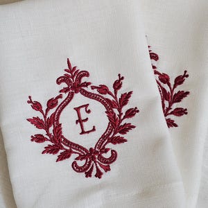 以下が含まれることがあります： 中央に深い赤色の刺繍で「E」のモノグラムが施された、オフホワイトのリネンのナプキン2枚。文字は装飾的で左右対称の植物のフレームに囲まれており、細部まで丁寧にステッチが施されたクラシックで構造的なデザインが特徴です。