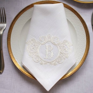 May include: A white linen napkin with a gold embroidered monogram of the letter "B" in a decorative frame. The napkin is folded and placed on a white plate with a gold rim.