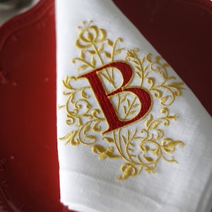 May include: White linen napkin with a gold embroidered floral design and a red letter "B". The napkin is folded and placed on a red plate. The design is ornate and elegant, suitable for formal dining.