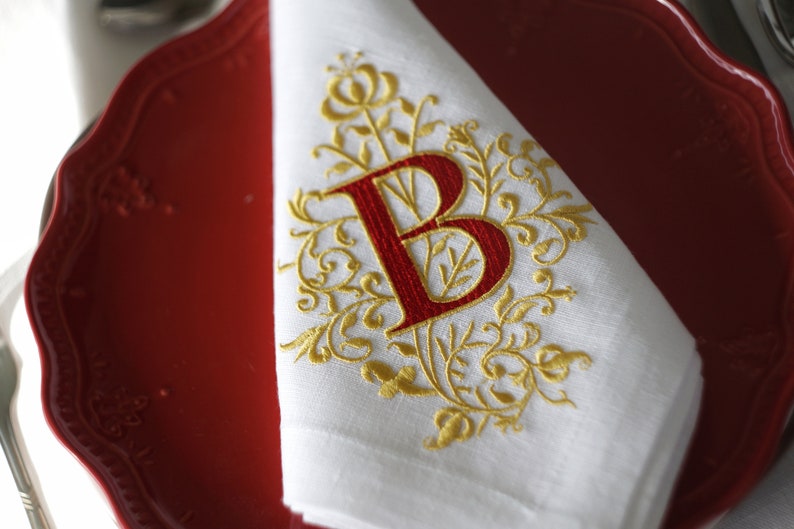 May include: White linen napkin with a gold embroidered floral design and a red letter "B". The napkin is folded and placed on a red plate. The design is ornate and elegant, suitable for formal dining.