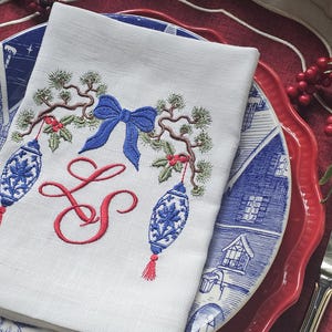 May include: A white cloth napkin featuring an embroidered design of a blue bow, red monogrammed letters LS, and two hanging blue and white ornaments suspended from green pine branches with small red berries, centred on a dinner plate.