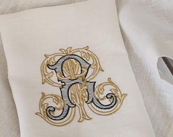 May include: A white linen napkin featuring an embroidered monogram in the centre. The design consists of a large, ornate letter R stitched in black thread with fine horizontal lines, surrounded by intricate gold-coloured decorative scrollwork.