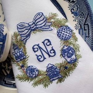 May include: White linen napkin with a blue and green embroidered Christmas wreath design. The wreath features a bow, baubles, and a monogram "N".