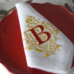 May include: White linen napkin with a red embroidered letter "B" and gold floral design, placed on a red plate. The napkin is folded and ready for use, with silverware and a white tablecloth in the background.