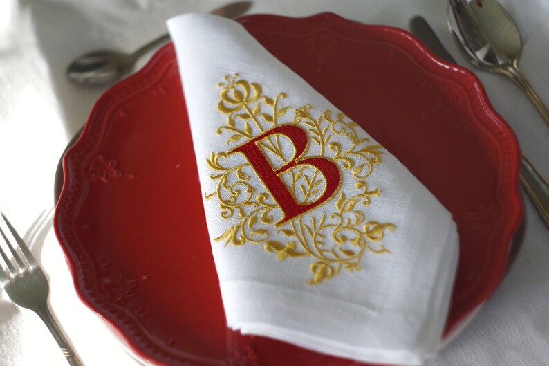 May include: White linen napkin with a red embroidered letter "B" and gold floral design, placed on a red plate. The napkin is folded and ready for use, with silverware and a white tablecloth in the background.