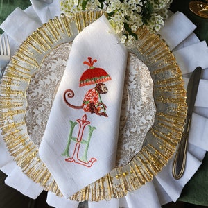 Peut inclure: Serviette en lin blanc avec broderie dorée d'un singe tenant un parapluie et la lettre "H". La serviette est pliée et placée sur une assiette à motifs dorés et blancs.