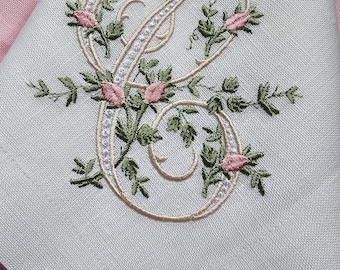 May include: A light green linen napkin with an embroidered letter "C". The letter is embellished with a floral design of pink roses and green leaves. The napkin is folded, with a pink background.