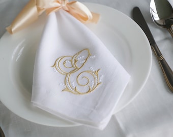 May include: A white linen napkin with a gold embroidered letter 'S' in a decorative script. The napkin is folded and placed on a white plate with a peach-coloured satin ribbon.