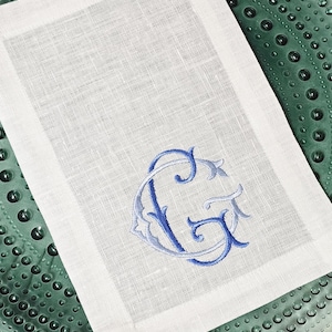 May include: White linen napkin with a blue embroidered monogram. The monogram features the letters "G" and "C" in a decorative, flowing script. The napkin is rectangular with a simple hem.