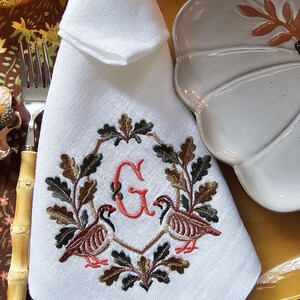 May include: A white cloth napkin featuring an embroidered design of two game birds facing each other, surrounded by an oak leaf wreath and a central red letter G. The embroidery uses shades of brown, green, and red thread.
