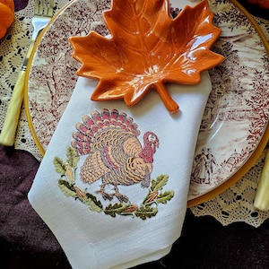 May include: A white cloth napkin featuring an embroidered turkey surrounded by green oak leaves and acorns. An orange ceramic dish shaped like a maple leaf rests on top of the napkin, which is placed on a plate with a brown and white landscape scene.