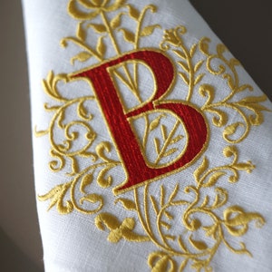 May include: White linen napkin with a red embroidered letter "B" and gold floral embellishments. The embroidery features intricate details and a classic design, suitable for formal dining or special occasions.