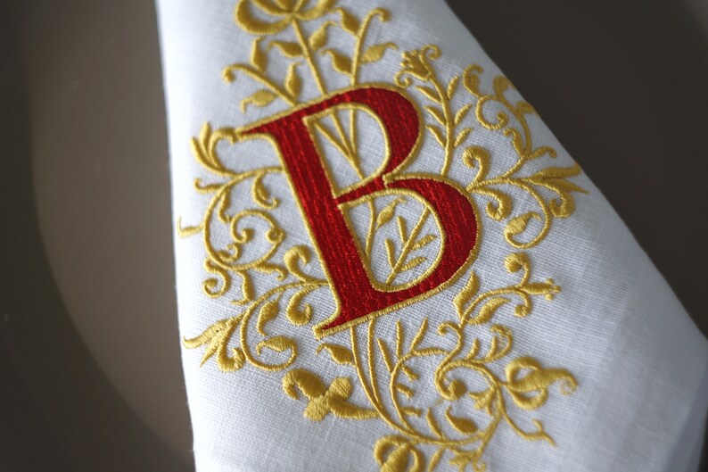 May include: White linen napkin with a red embroidered letter "B" and gold floral embellishments. The embroidery features intricate details and a classic design, suitable for formal dining or special occasions.