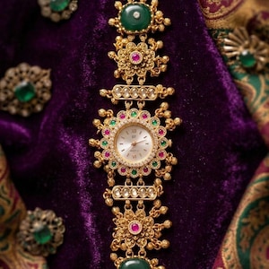 May include: An ornate gold-toned wristwatch with a white watch face and gold hands. The watch band is adorned with green and red gemstones and floral designs. The watch is set against a purple velvet background.