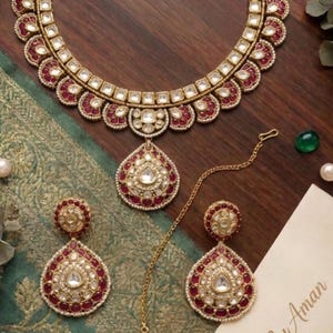 May include: A gold-toned jewelry set featuring a necklace, two teardrop-shaped earrings, and a matching headpiece. Each piece is adorned with clear, square-cut stones and deep red accents. A card with the text SequinsByAman is visible nearby.