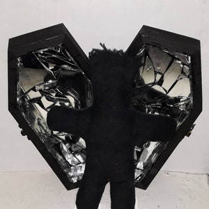 May include: A black fabric voodoo doll standing in front of a black wooden heart-shaped box with a broken mirror inside.