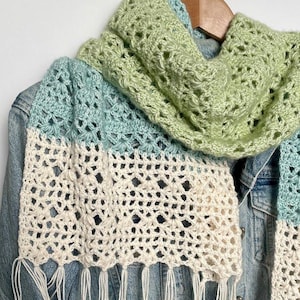 Könnte beinhalten: Handgefertigter Häkelschal in Grün-, Blau- und Cremetönen. Der Schal hat einen Fransenrand und ist über einer Jeansjacke drapiert. Das offene Webmuster ist im gesamten Schal sichtbar.