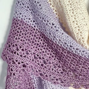 May include: A handmade crochet shawl in a gradient of colors, transitioning from cream to lavender and deep purple. The openwork design creates a delicate, lacy texture. The shawl is draped, showcasing its intricate stitchwork and color variations.