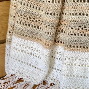 May include: A cream, beige, and brown crochet blanket with an openwork design. The blanket has a white fringe along the bottom. The soft, textured blanket is ideal for home decor and measures approximately 150cm x 180cm.