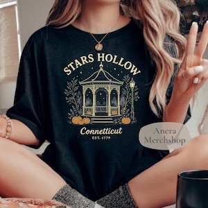 Könnte beinhalten: Schwarzes T-Shirt mit einem goldenen und weißen Grafikdesign eines Pavillons, Kürbissen und dem Text "Stars Hollow Connecticut EST. 1779". Das T-Shirt wird von einer Person getragen, die mit gekreuzten Beinen sitzt.