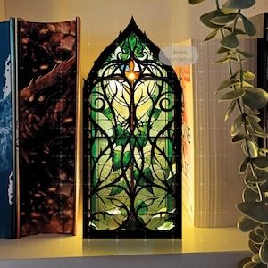 Könnte beinhalten: Eine dekorative Lampe in Form eines Buntglasfensters mit schwarzem Rahmen und grünem und gelbem Glas. Die Lampe hat ein detailliertes Design aus Blättern und Ranken. Die Lampe steht auf einem Regal mit Büchern und einer Pflanze.