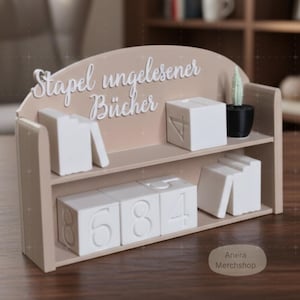 Op de afbeelding: Een beige houten plank met de Duitse tekst "Stapel ungelesener Bücher" in witte letters. Het bevat witte genummerde blokken en kleine boekvormige objecten. Een kleine cactus in een zwarte pot staat er ook op.