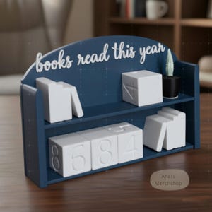 Könnte beinhalten: Ein marineblaues Bücherregal-Schreibtisch-Organizer mit dem Text "books read this year" in weißer Schrift. Es verfügt über weiße Zahlenblöcke und buchförmige Akzente sowie einen kleinen Topf-Kaktus.
