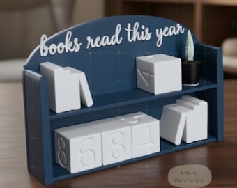 May include: A navy blue bookshelf-shaped desk organizer with the text "books read this year" in white script. It features white number blocks and book-shaped accents, along with a small potted cactus.