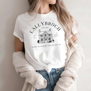 May include: White t-shirt with a black graphic design of a house with the text "Lallybroch Est 1702" above the house and "Take me home" to the left of the house and "To Lallybroch" to the right of the house and "Home is where the heart is" below the house.