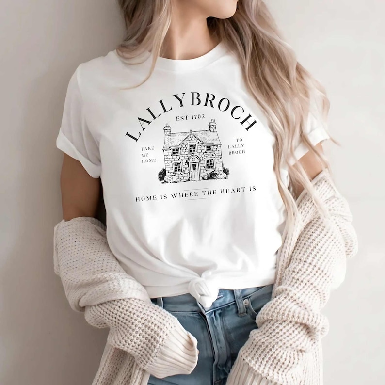 May include: White t-shirt with a black graphic design of a house with the text "Lallybroch Est 1702" above the house and "Take me home" to the left of the house and "To Lallybroch" to the right of the house and "Home is where the heart is" below the house.