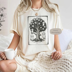 May include: Cream-colored t-shirt featuring a black and white illustration of a tree with four small, ghost-like figures. The tree has detailed leaves and roots. The text "Anera Merchshop" is printed on the shirt.