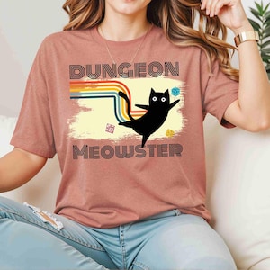 Può includere: Una maglietta rosa con un design arcobaleno retrò e un gatto nero con il testo "Dungeon Meowster".
