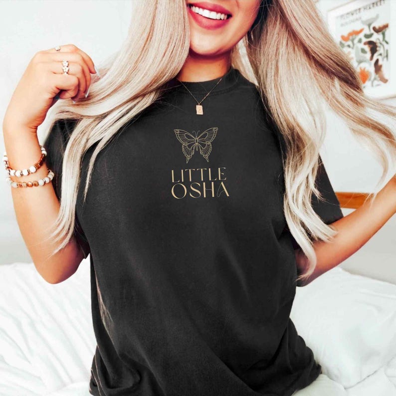 May include: Black t-shirt with a gold butterfly graphic and the text "LITTLE OSHA" printed on the front.