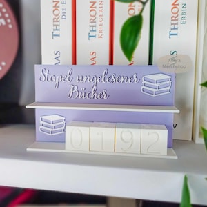 Può includere: Un espositore per libri lilla con la scritta "Stapel ungelesener Bücher" e una grafica di pila di libri. L'espositore ha un ripiano e un display della data con i numeri 0192.