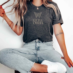 May include: A dark gray t-shirt with a gold butterfly graphic and the text "LITTLE OSHA" printed on the front.